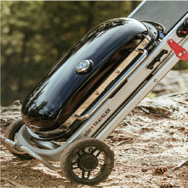 Portable charcoal grill with a sleek black lid mounted on a wheeled cart, positioned outdoors on a natural surface.