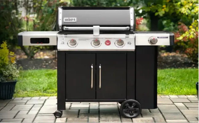 Black outdoor grill with stainless steel side shelves and control knobs, set on a stone patio with green shrubbery in the background.