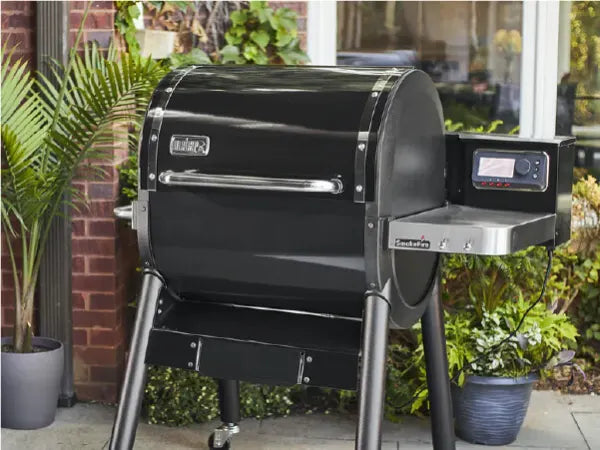Large black outdoor pellet grill with a digital control panel and stainless steel side shelf, set on a patio with surrounding plants and brick wall.