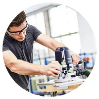 Man wearing safety glasses and a dark t-shirt operating a power woodworking tool on a piece of wood in a bright workshop.