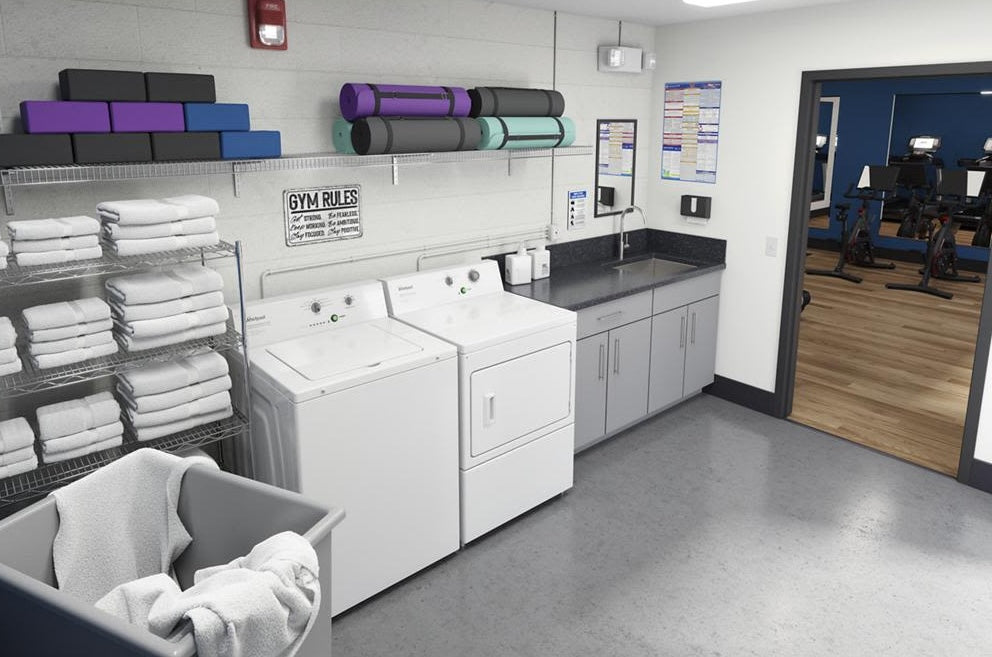 White commercial electric dryer and matching washer in gym laundry room with neatly stacked towels and yoga mats nearby