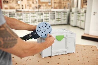 Person attaching blue abrasive disc to power sander in workshop with organized Festool tool storage box nearby