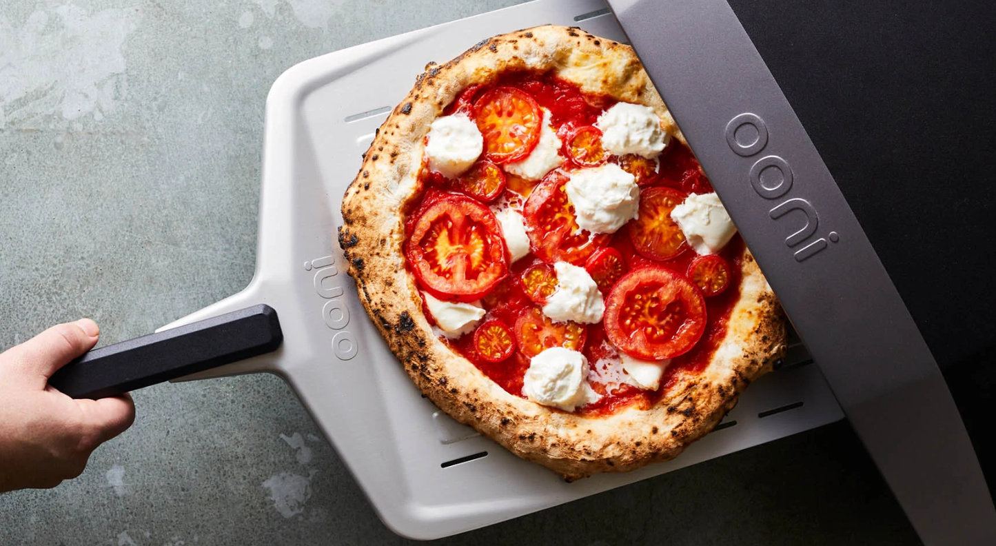 Freshly baked pizza with tomato slices and dollops of cheese being removed from a black and silver ooni pizza oven in outdoor living grilling setup