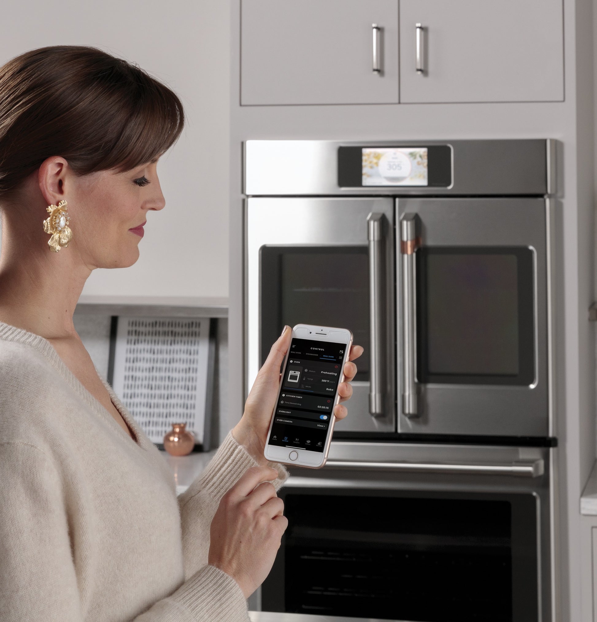 Woman using a smartphone app to control a stainless steel built-in convection French-door wall oven in kitchen