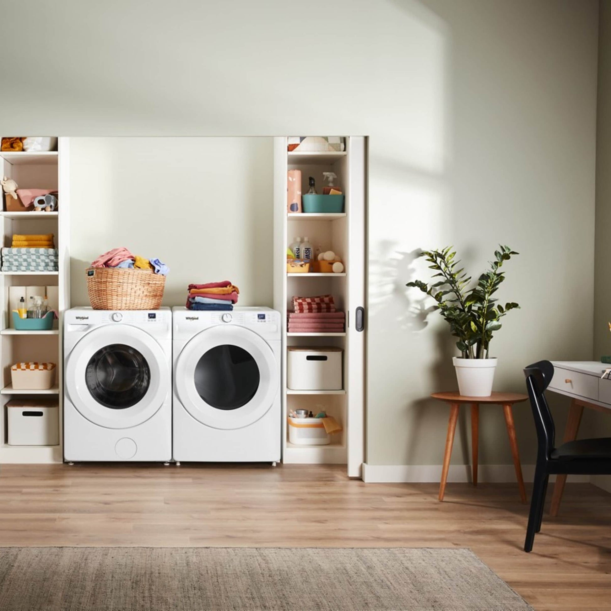 Compact laundry area with white front load washer and dryer, built-in shelving, plants, and organized household items nearby