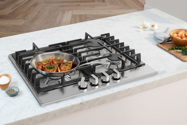 Stainless steel gas cooktop with four burners and heavy-duty cast iron grates cooking seasoned meat on marble countertop