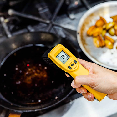 Hand holding yellow infrared temperature gun measuring heat from cast iron pan on stove in grilling setting