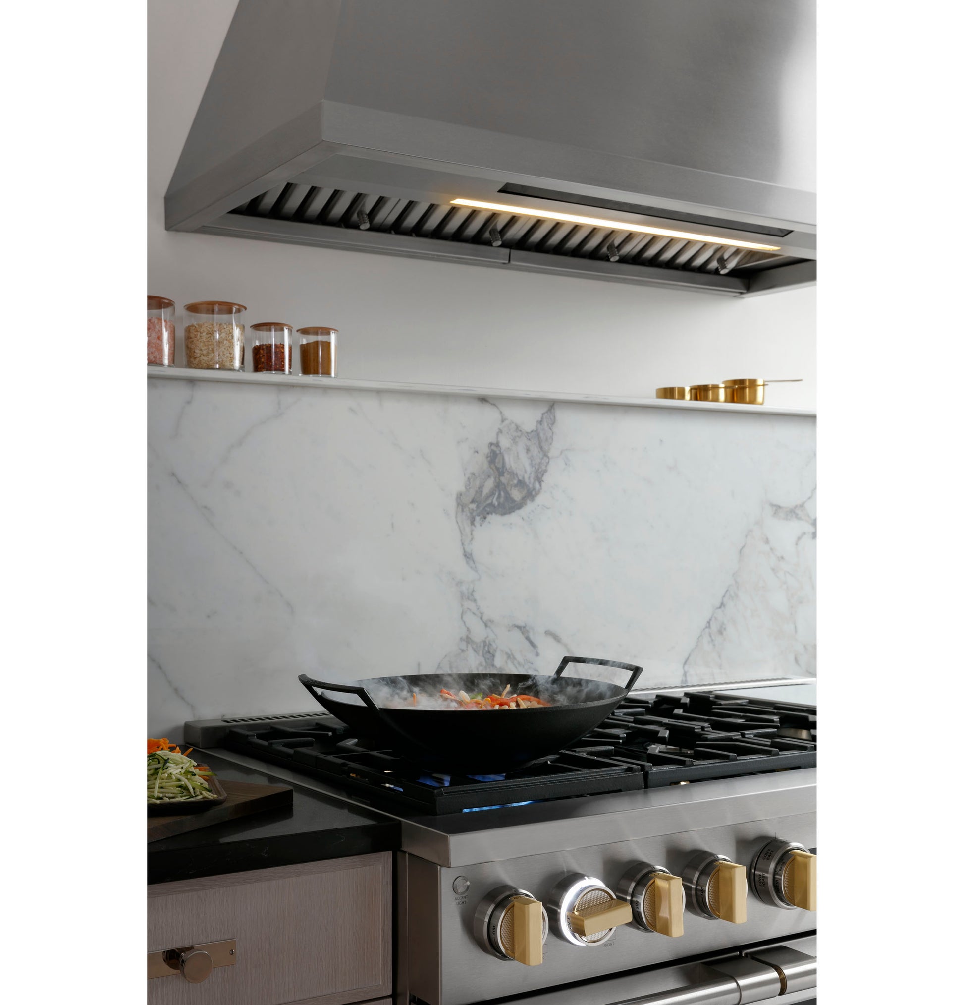 Stainless steel professional kitchen hood above gas cooktop with black wok and marble backsplash in appliances collection