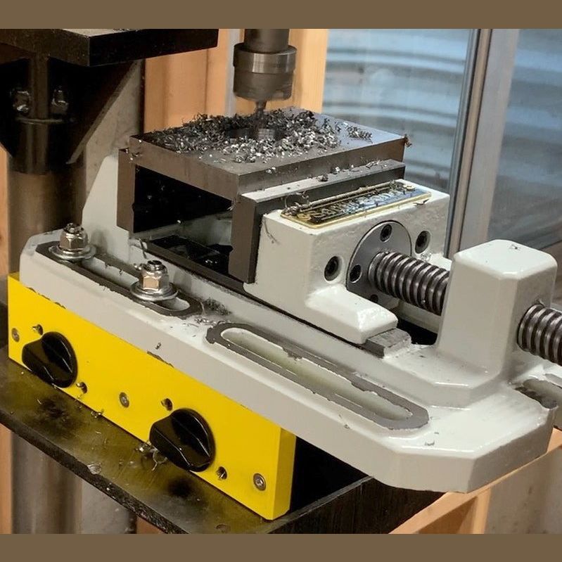Metal drill press vise mounted on a yellow and white base with metal shavings in a tool accessories setting