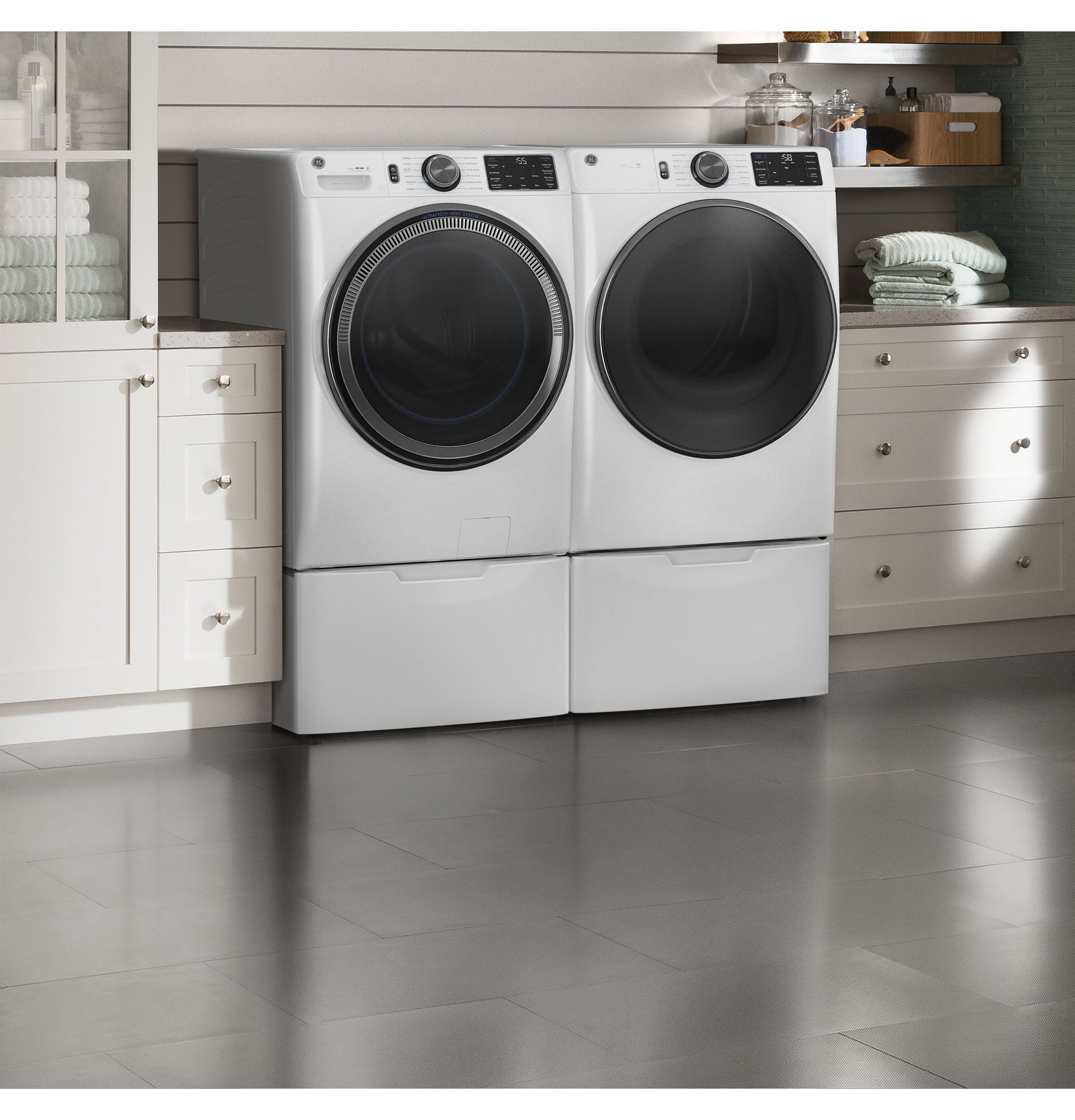 Smart front load white washer and dryer pair with pedestal drawers in modern laundry room with shelves and towels