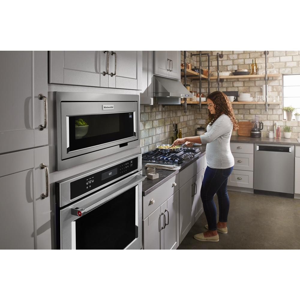 Woman cooking in modern kitchen with stainless steel appliances, gray cabinetry, brick backsplash, and under-cabinet ventilation hood
