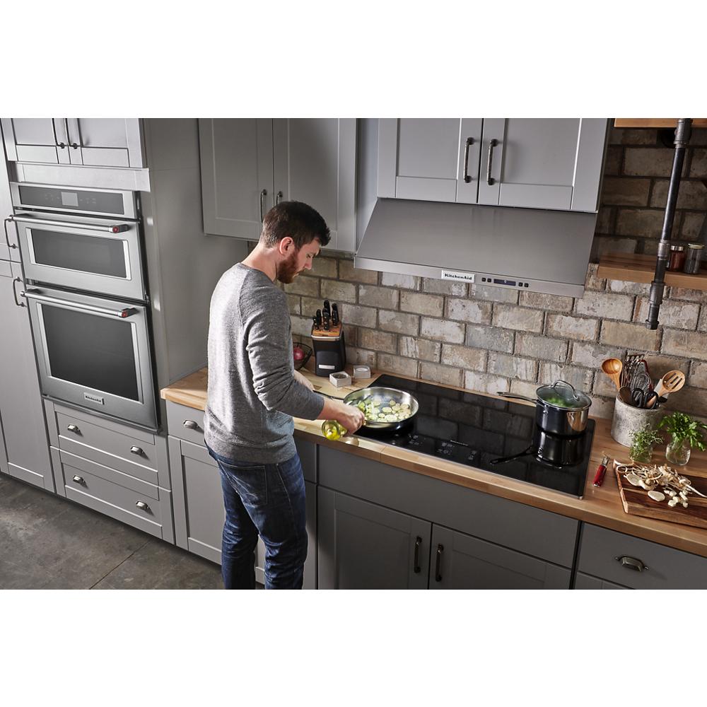 Man cooking with vegetables on a black glass electric cooktop in a modern kitchen with gray cabinetry and brick backsplash