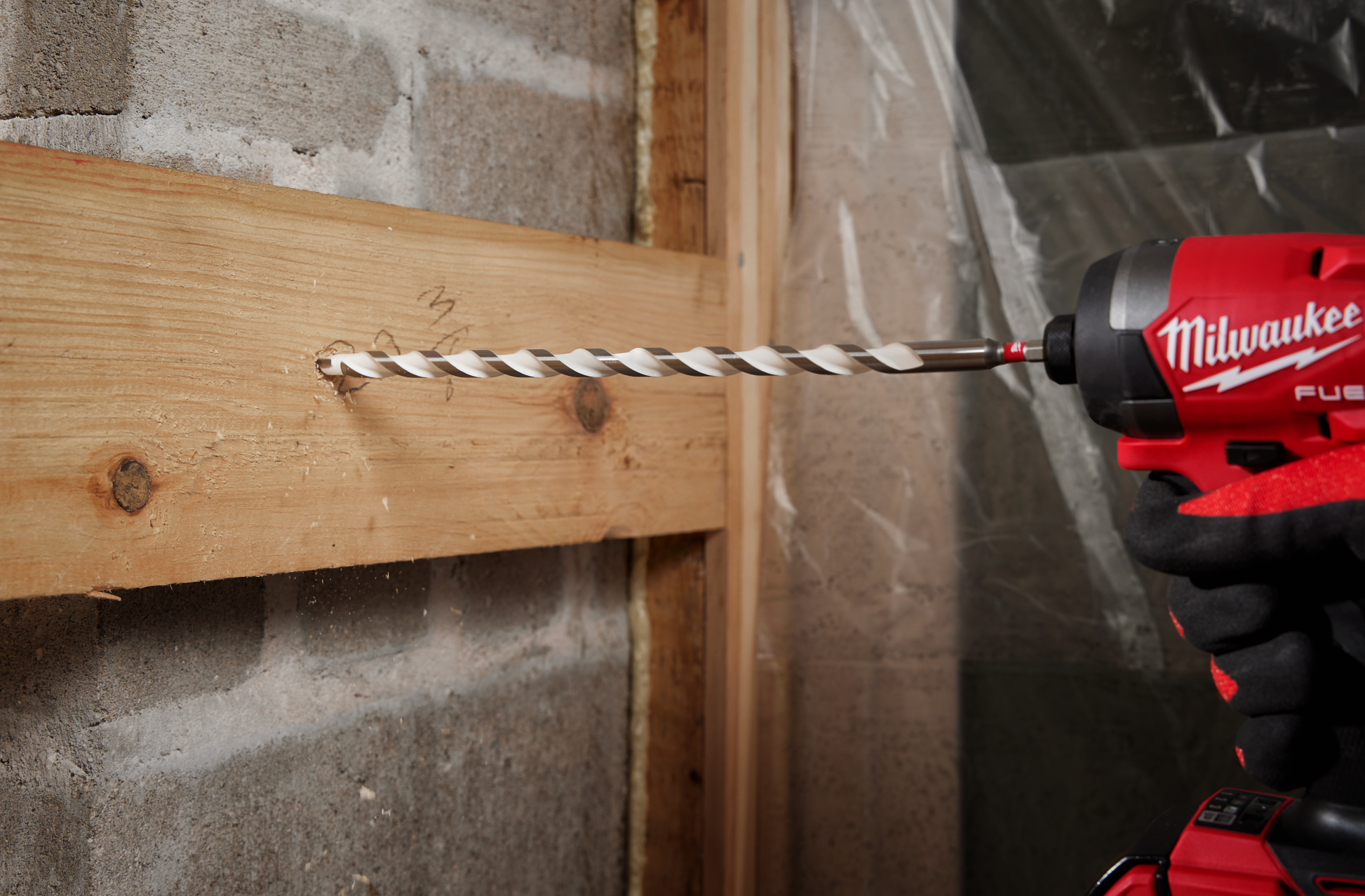 Red and black Milwaukee power drill with a multi-material bit drilling into a wooden beam in a construction setting