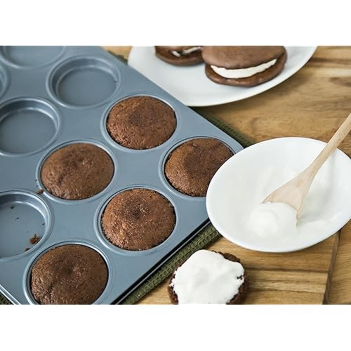 Gray non-stick pan with round molds baking chocolate whoopie pies beside bowl of white frosting in kitchenware collections