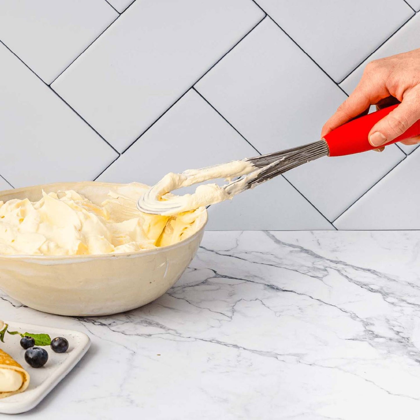 Hand using red-handled stainless steel whisk to scoop batter from beige mixing bowl on marble counter with tile background