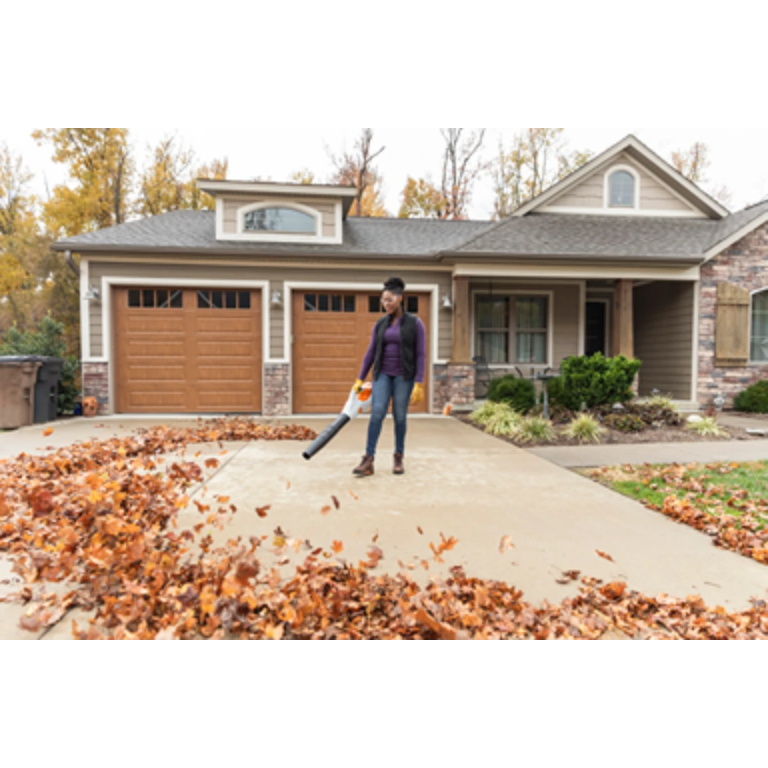 Person using cordless STIHL handheld blower to clear fallen autumn leaves from a residential driveway and sidewalk