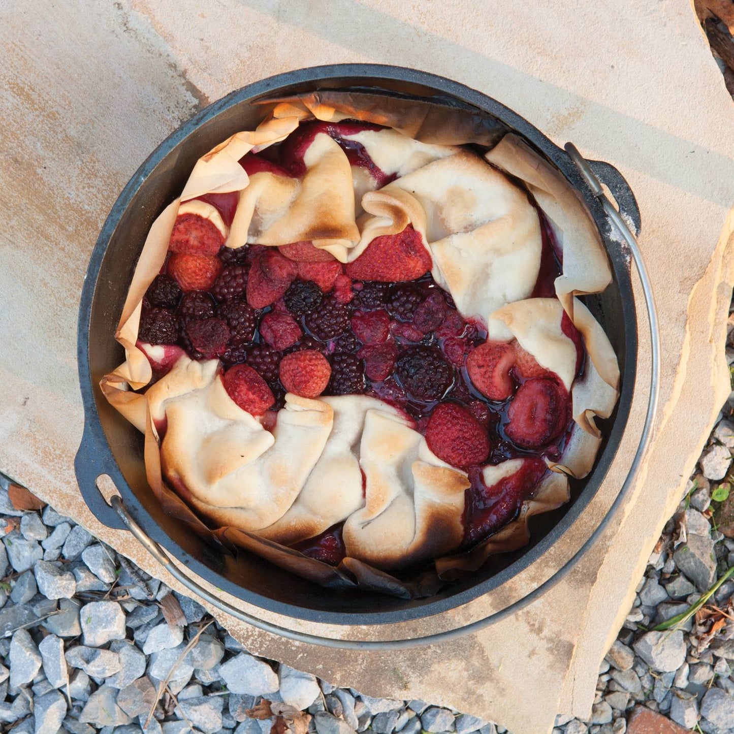 Cast iron Dutch oven with mixed berry cobbler baking outdoors on stone surface in kitchenware collection