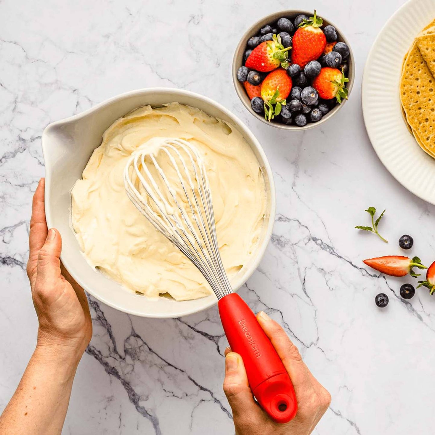 Hand holding red stainless steel whisk mixing batter in white ceramic bowl with fresh fruit and plate nearby in kitchenware setting