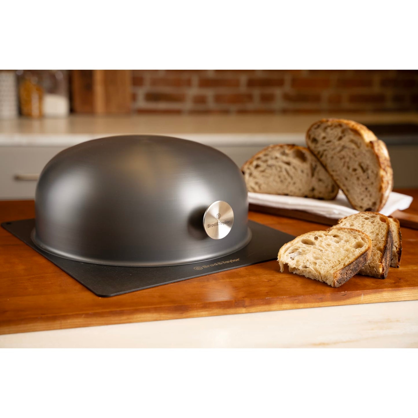 Matte black domed baking shell with stainless steel logo badge next to sliced artisanal bread on wooden countertop in kitchenware collection
