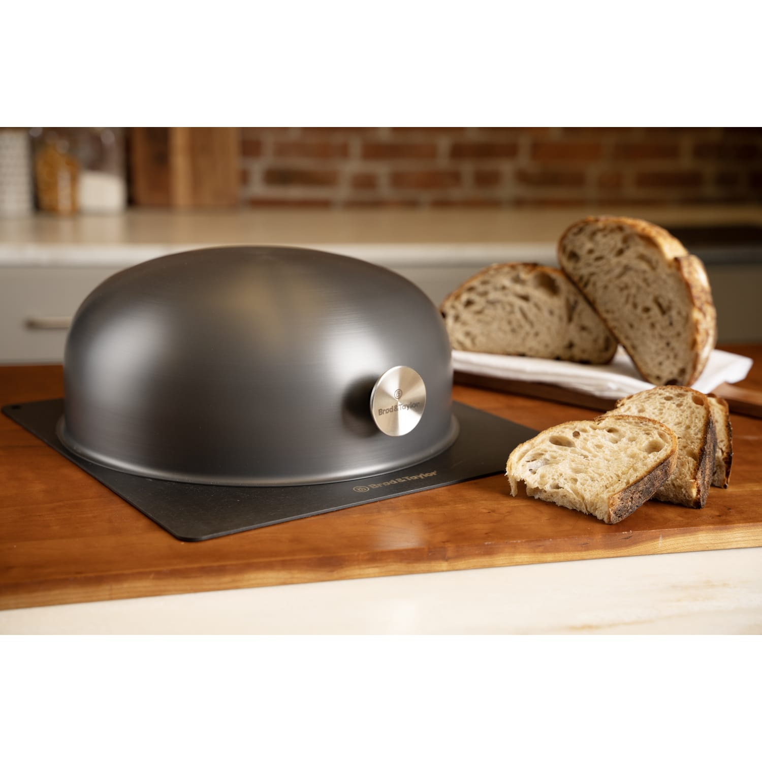 Matte black domed baking shell with stainless steel logo badge next to sliced artisanal bread on wooden countertop in kitchenware collection