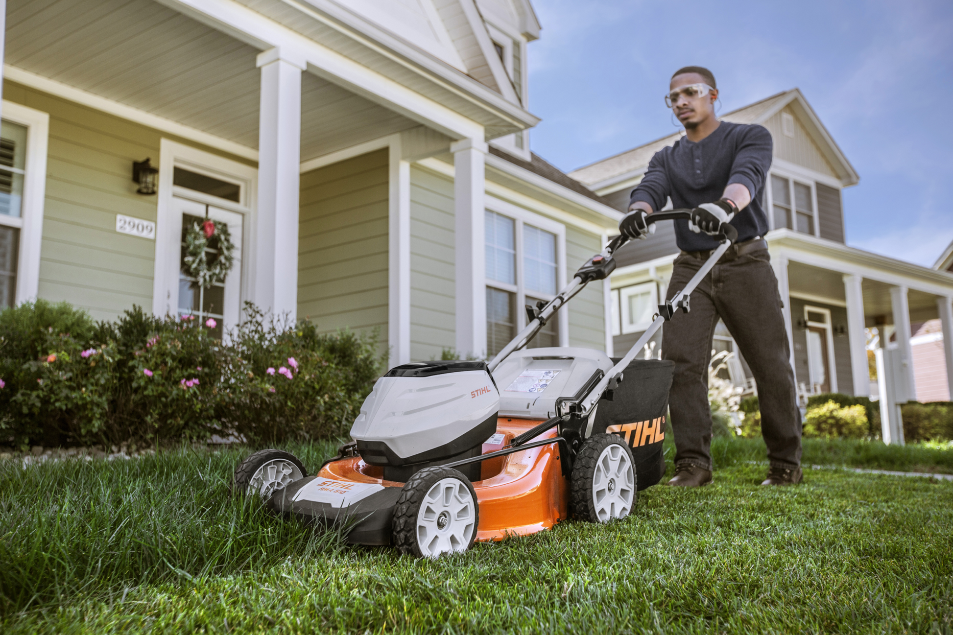 Person using a white and orange battery-powered STIHL lawn mower on green grass in front of a suburban house