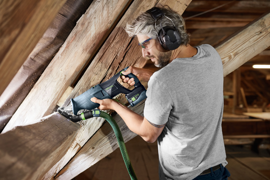 Man wearing safety glasses using a Festool cordless reciprocating saw on angled wooden beams in attic renovation