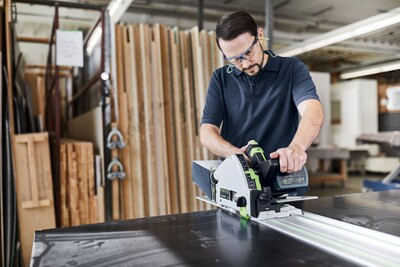 Man operating a power saw with a guide rail in a woodworking shop with wood panels in the background