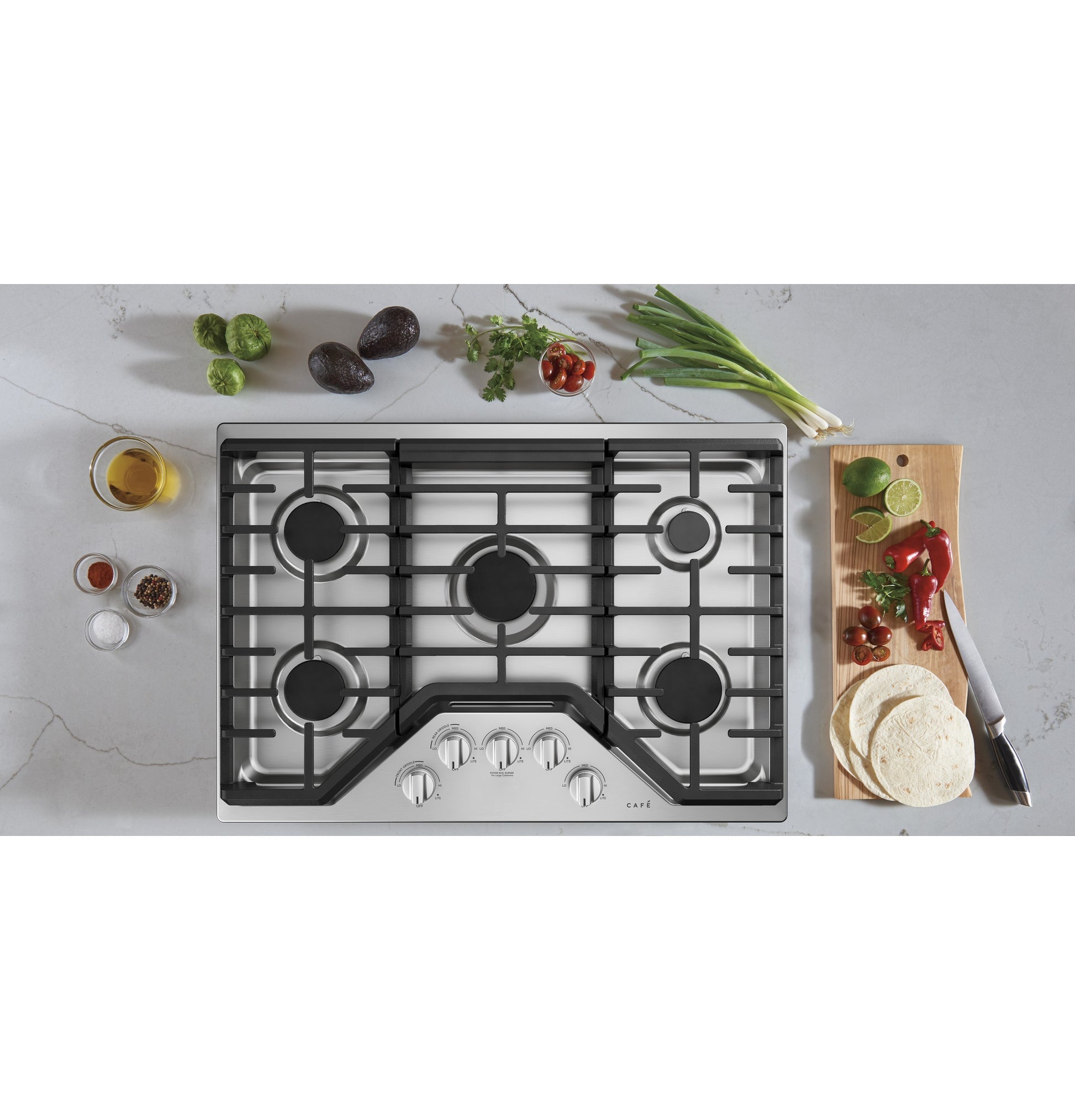 Stainless steel gas cooktop with five burners and control knobs surrounded by fresh ingredients for cooking from Gas Cooktops collection