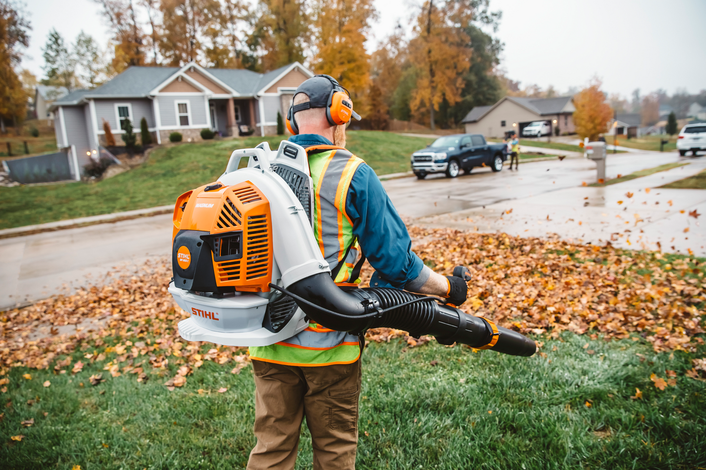 Person wearing protective gear using an orange backpack blower to clear fallen autumn leaves in a residential neighborhood