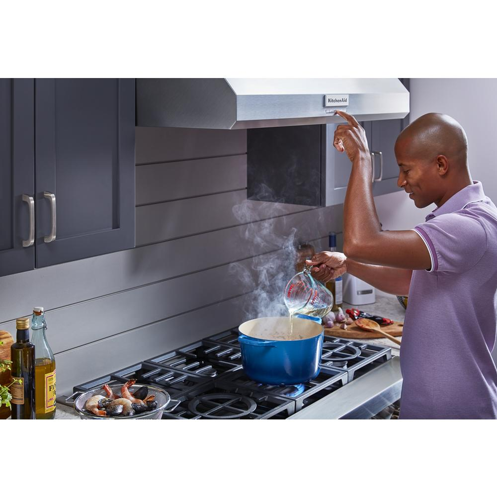 Man cooking with blue pot under stainless steel commercial-style wall-mount canopy range hood in kitchen setting