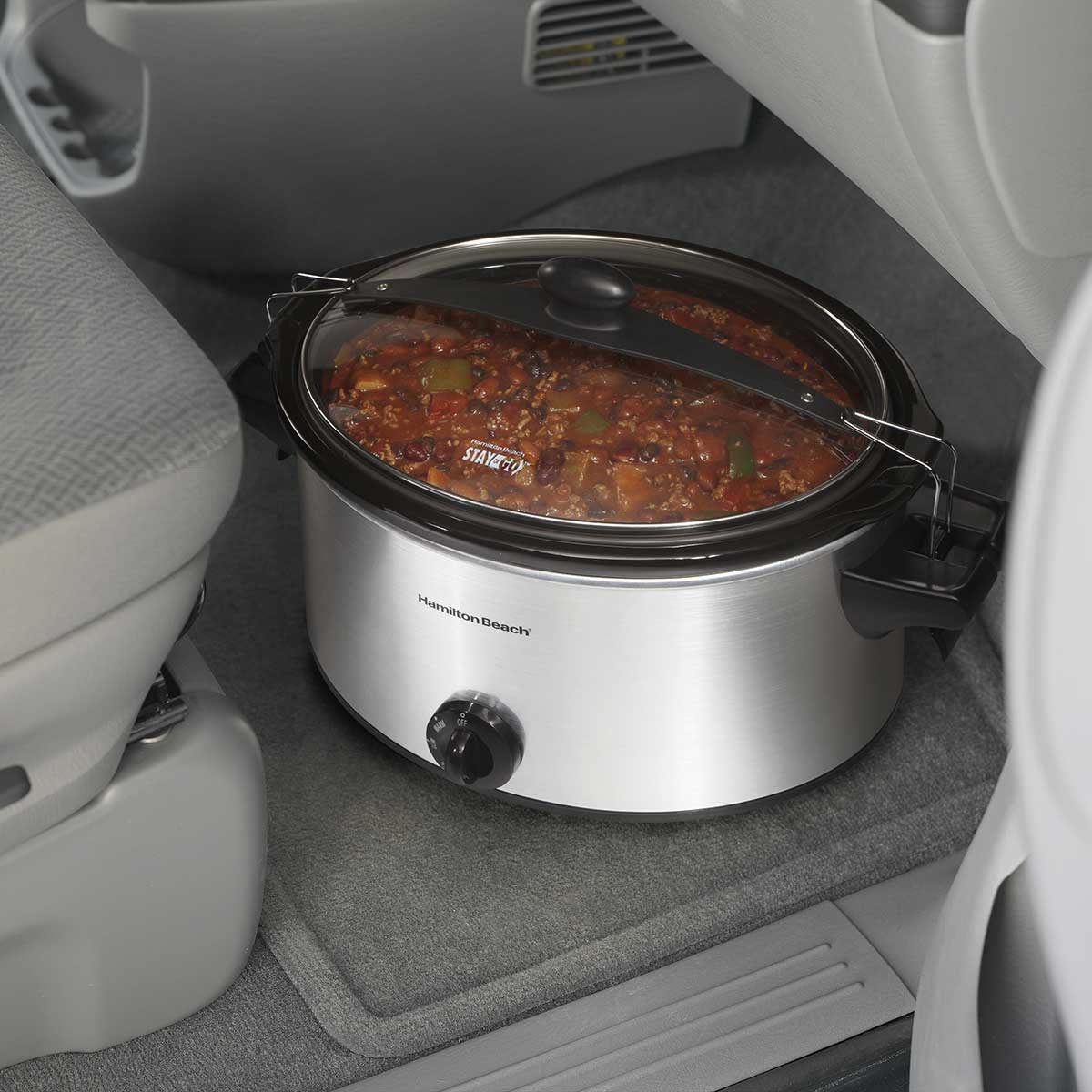 6-quart stainless steel slow cooker with clear locking lid placed on vehicle floor for portable cooking convenience