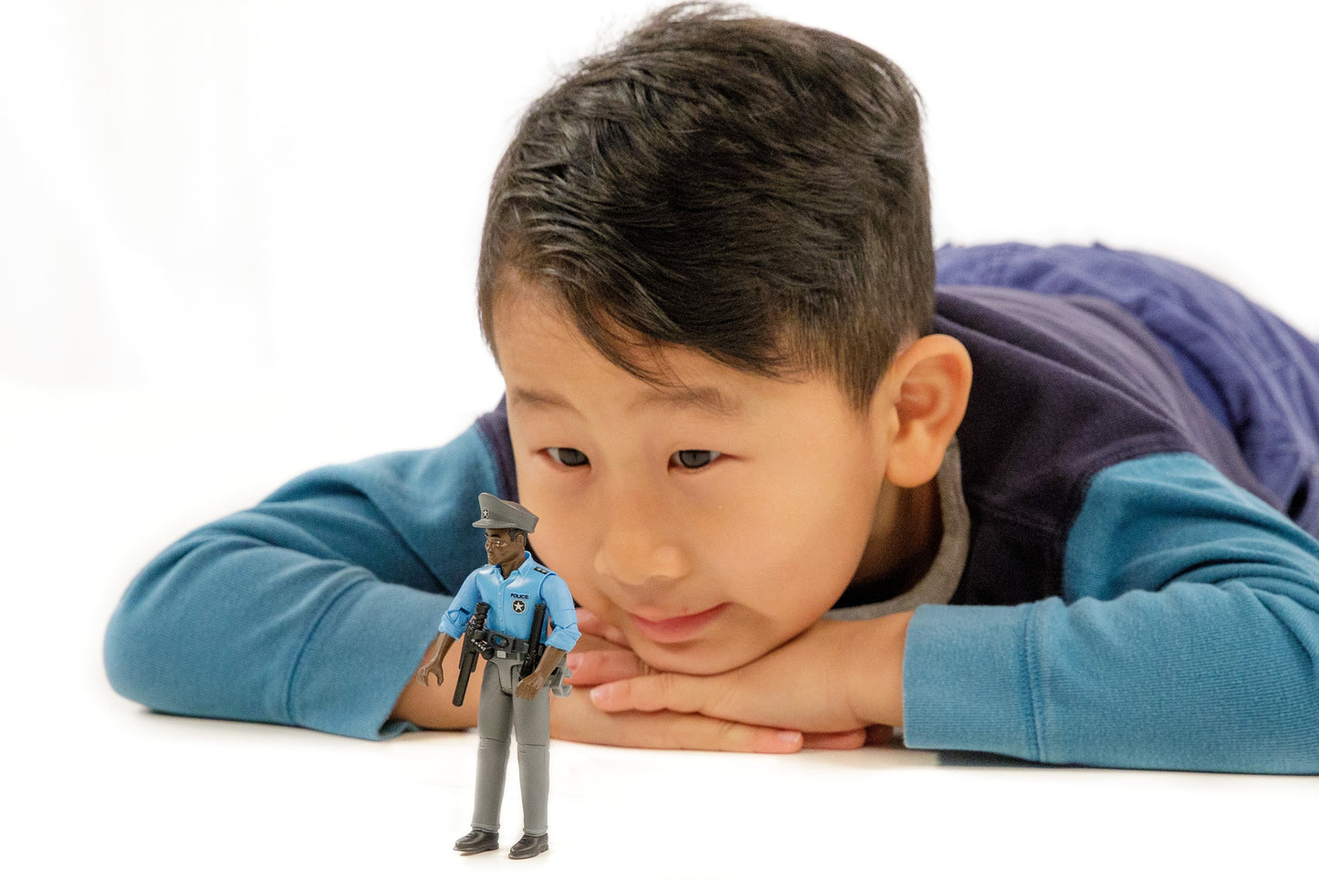 Young boy lying down looking intently at a small dark-skinned policeman toy figure with accessories