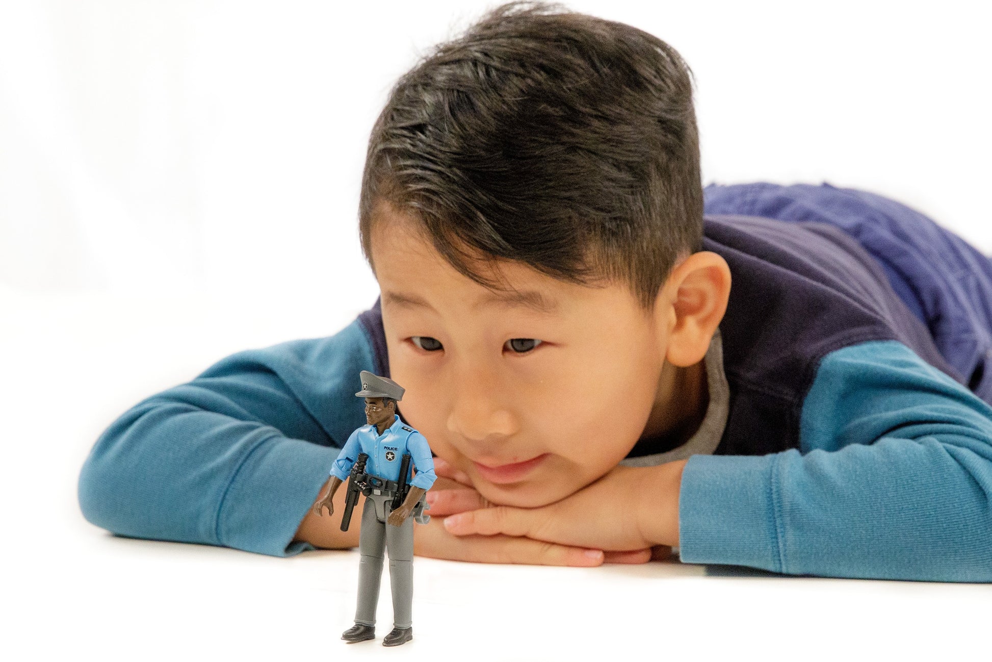 Young boy lying down looking intently at a small dark-skinned policeman toy figure with accessories