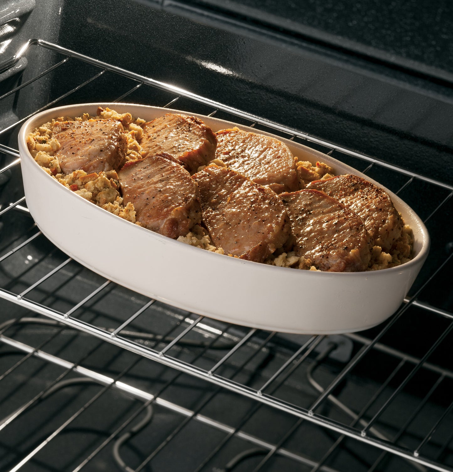 Six pork chops cooking on a white ceramic dish inside a free-standing oven in a home kitchen setting