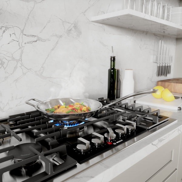 Stainless steel cooktop with control knobs, simmering vegetables in pan, kitchenware and cooking tools nearby