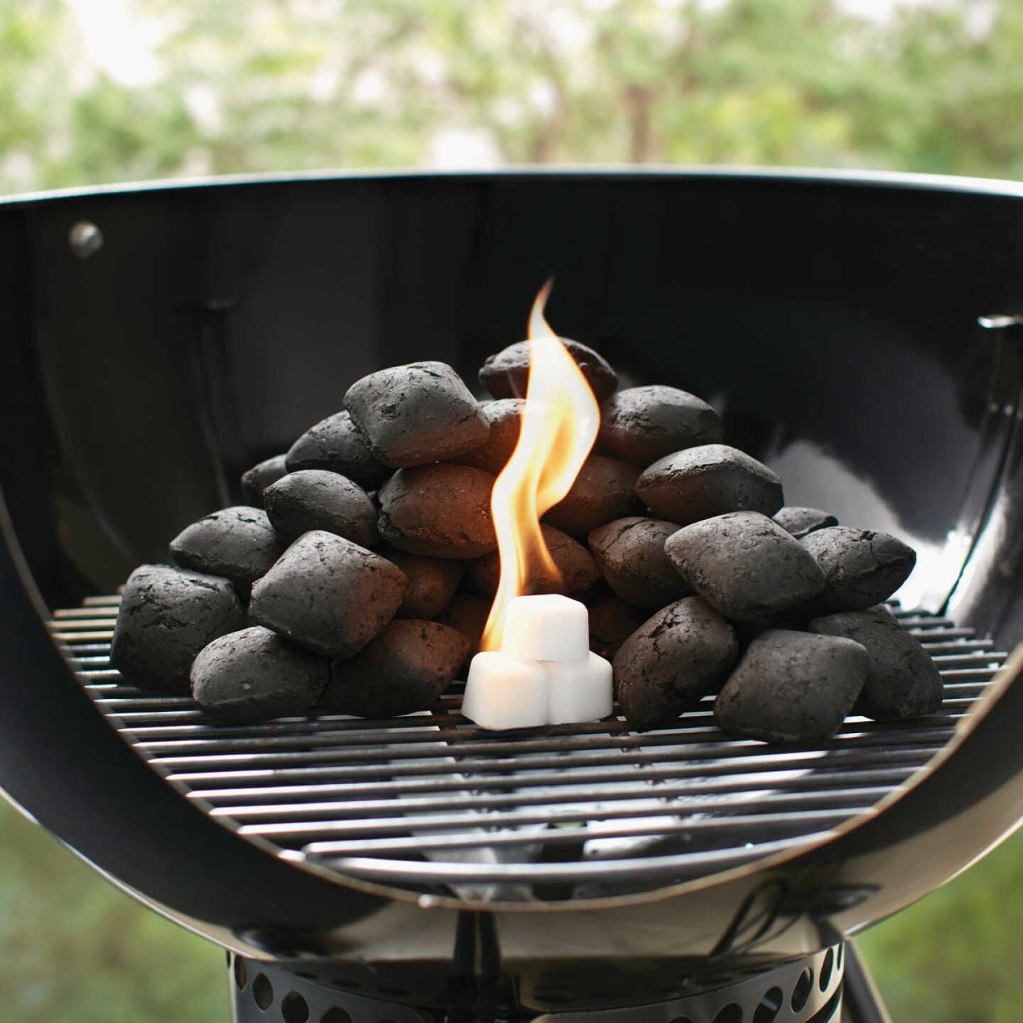 Flame igniting odorless starter cube among charcoal briquettes on round kettle grill for outdoor grilling.