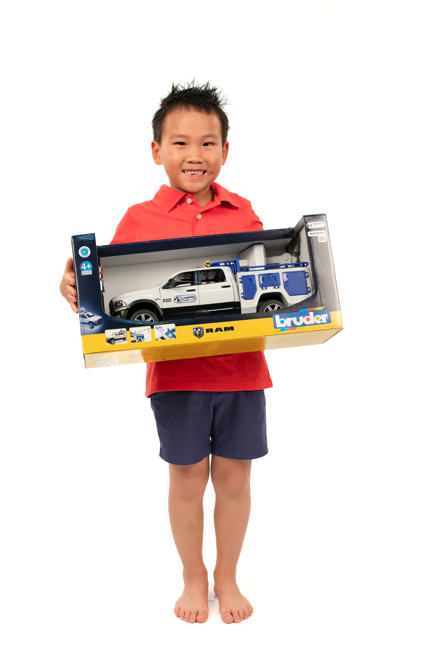 Smiling child in red shirt holding boxed Bruder RAM service truck toy with crane and rotating light for role play