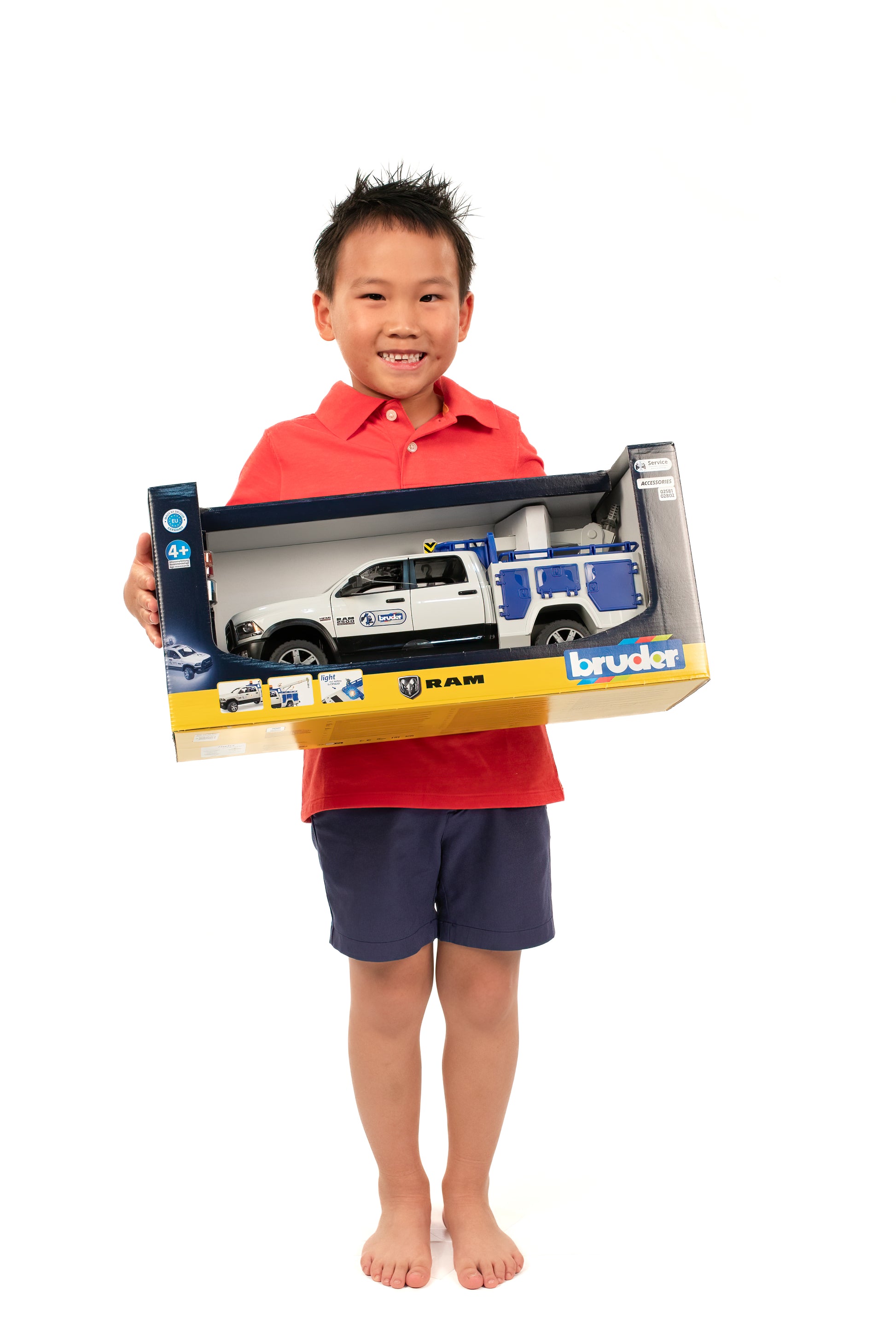 Smiling child in red shirt holding boxed Bruder RAM service truck toy with crane and rotating light for role play