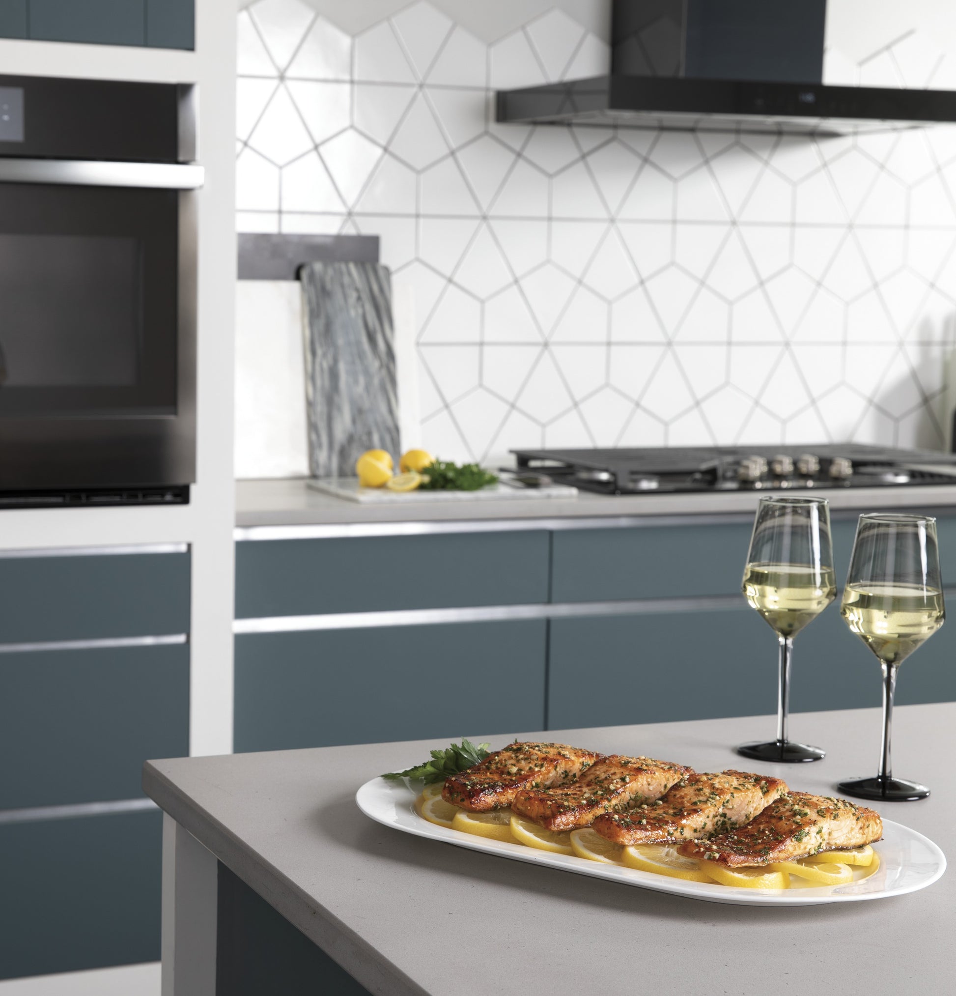 Modern kitchen with blue-gray cabinetry, built-in wall ovens, gas cooktop, and plated cooked salmon with wine glasses on island