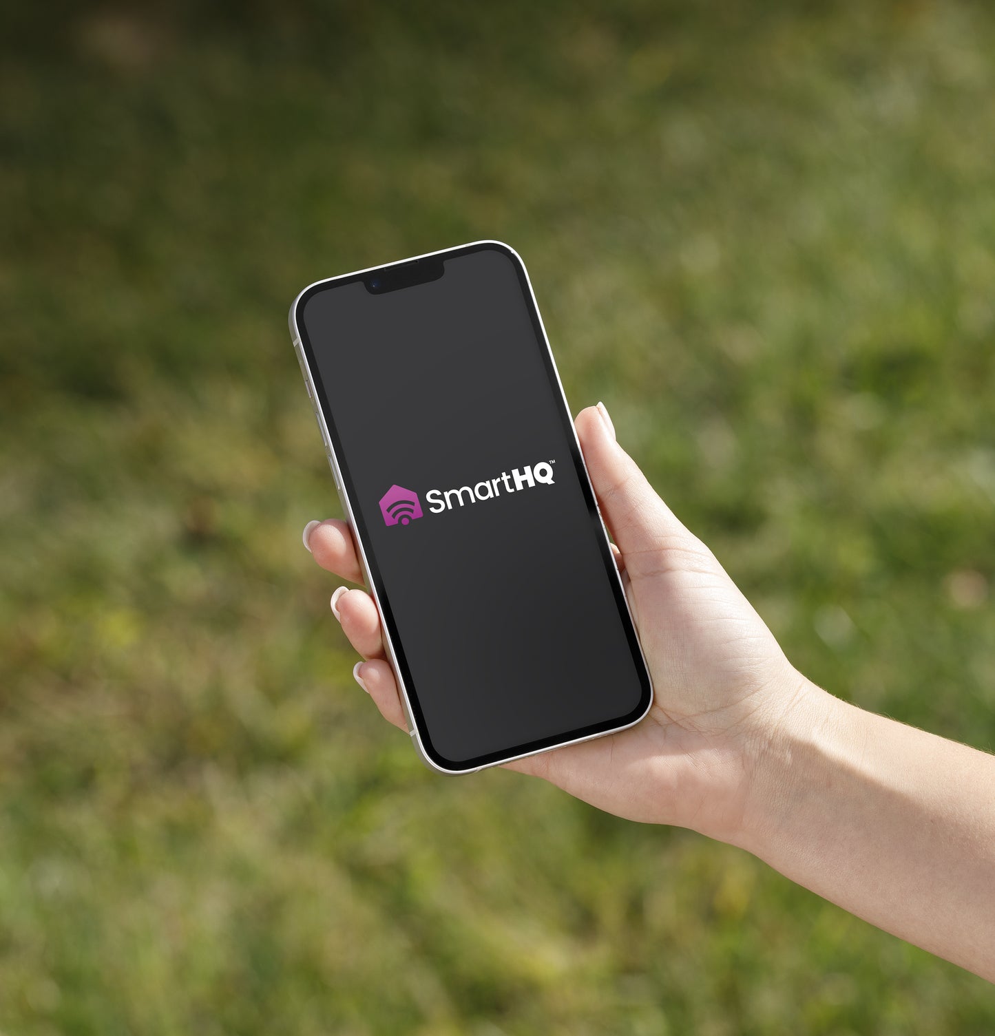 Hand holding smartphone displaying SmartHQ app logo on screen with blurred green outdoor background