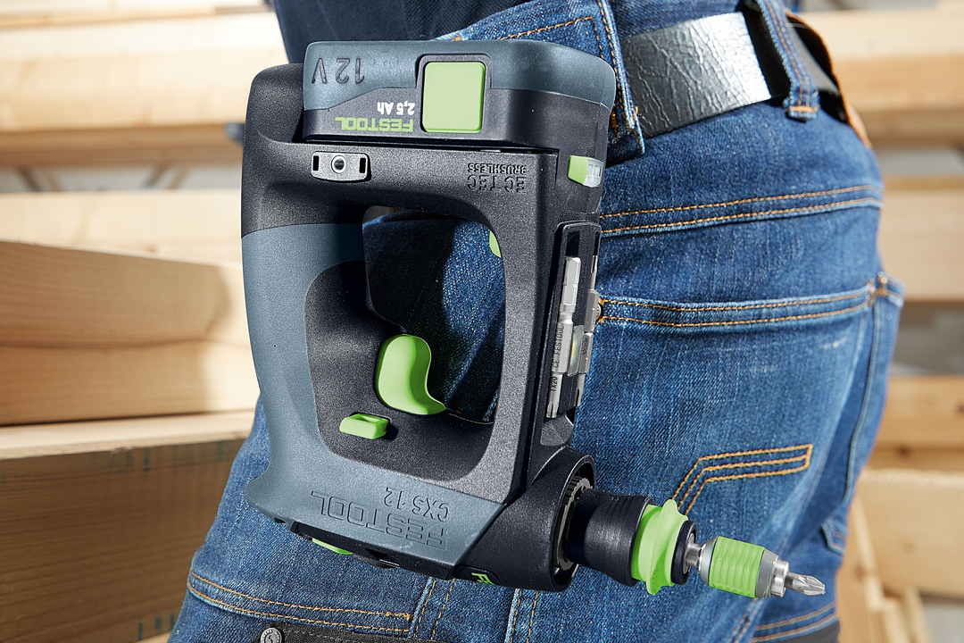 Compact cordless drill with ergonomic C-shaped grip clipped to blue jeans pocket on woodworking site