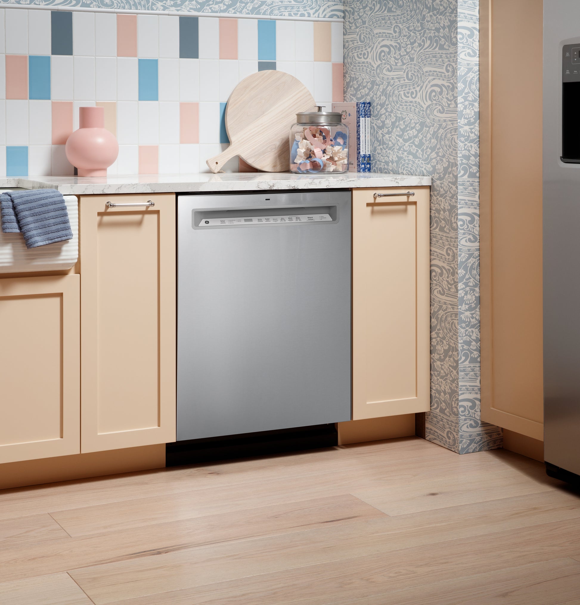 Stainless steel front control dishwasher installed under countertop in kitchen with pastel cabinets and patterned wall