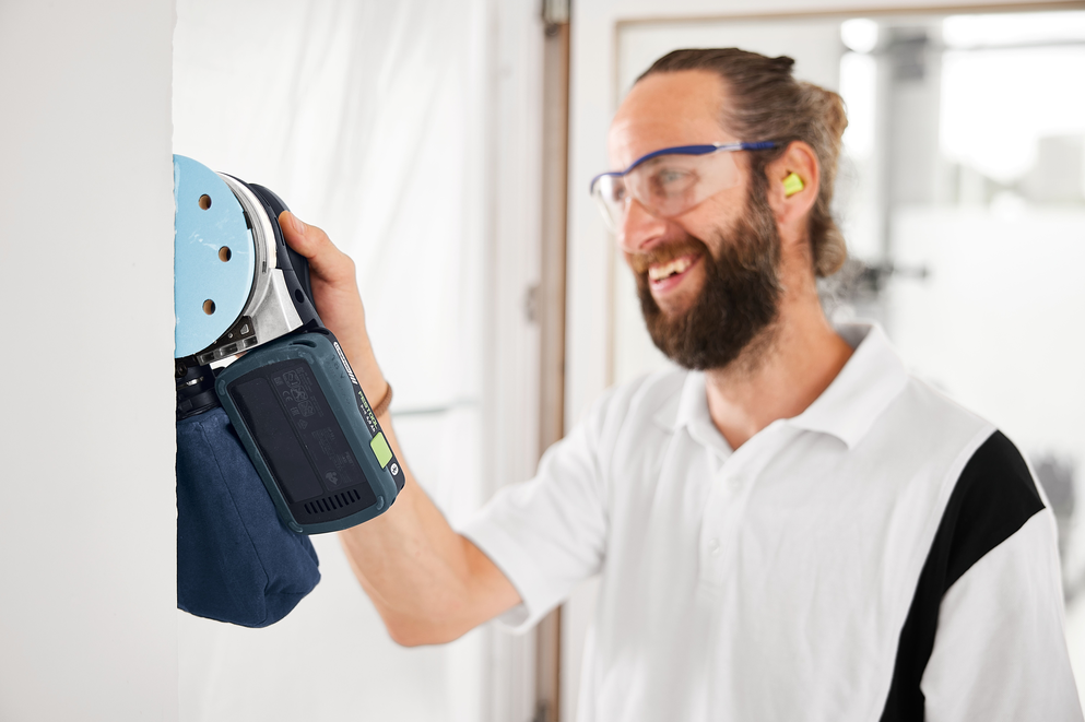 Bearded man wearing safety glasses and earplugs using cordless Festool random orbit sander on white wall in workshop