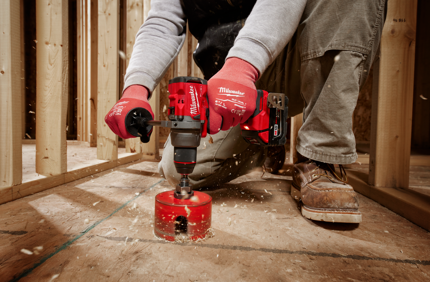 Person using red Milwaukee cordless drill with hole saw bit for wood drilling in framed wooden construction site