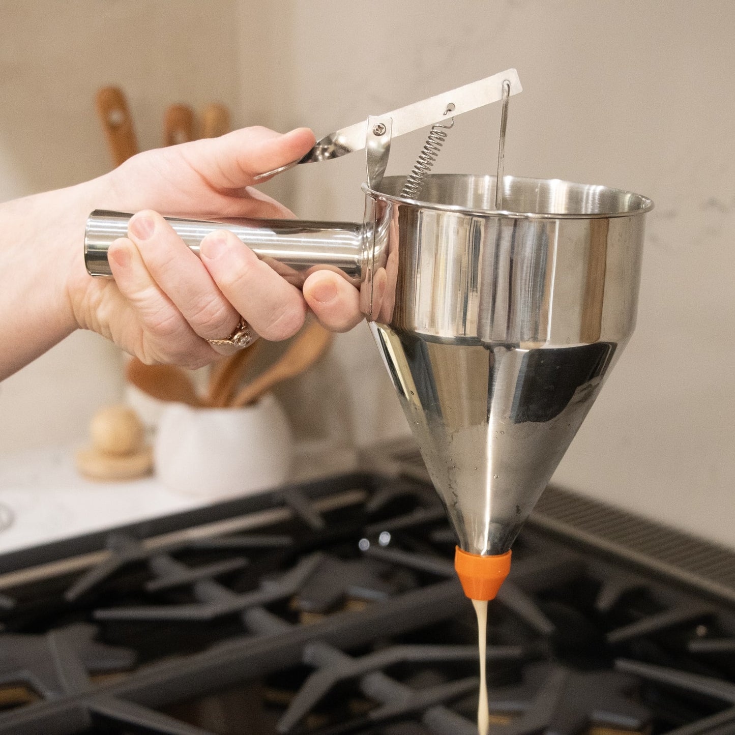 Hand holding stainless steel batter dispenser with orange silicone tip over stovetop in kitchenware setting