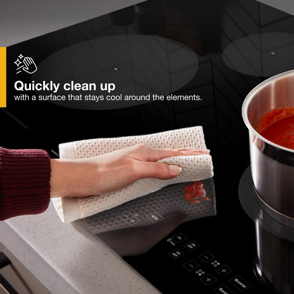 Hand wiping spill on black glass induction cooktop with touch controls and stainless steel pot simmering sauce nearby