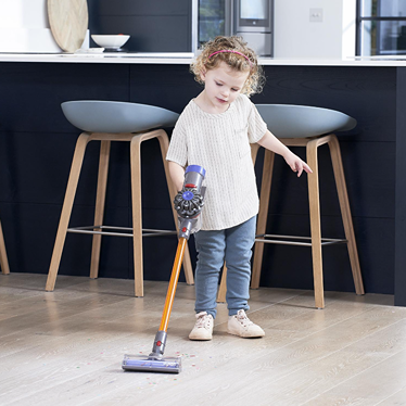 Young child using a colorful cordless vacuum toy to clean a hardwood floor in a modern kitchen setting