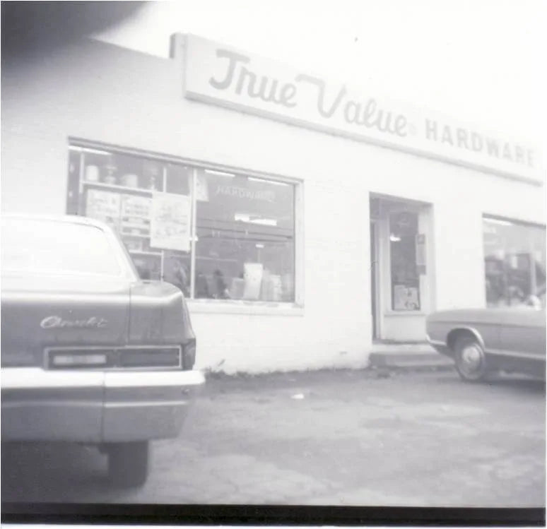 Black and white photo of a vintage hardware store with two classic cars parked in front, showcasing a simple storefront with large windows and a sign reading 
