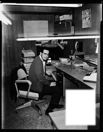 Black and white photo of a man wearing glasses and a suit seated in a mid-century office chair at a desk cluttered with papers and office supplies, in a wood-paneled room.