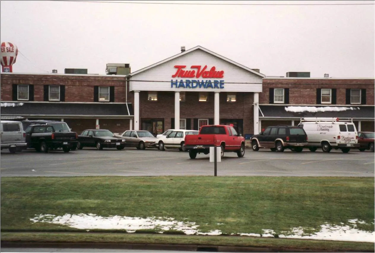 Two-story brick building with a white triangular entrance roof displaying red and blue signage for a hardware store, with several vehicles parked in front on an overcast day